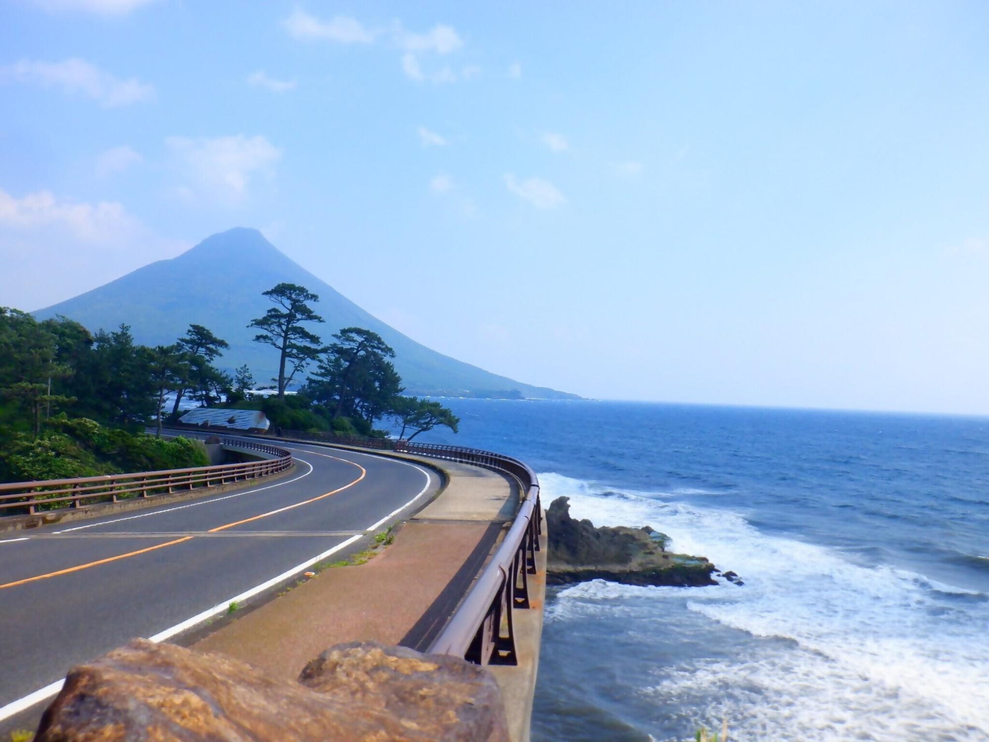 道路の右側に海が広がり、道路の先には開聞岳が見えている風景写真