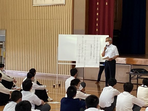 ホワイトボードに資料を張り出し、先生が話しているのを、体育座りをして聞いている生徒たちの写真