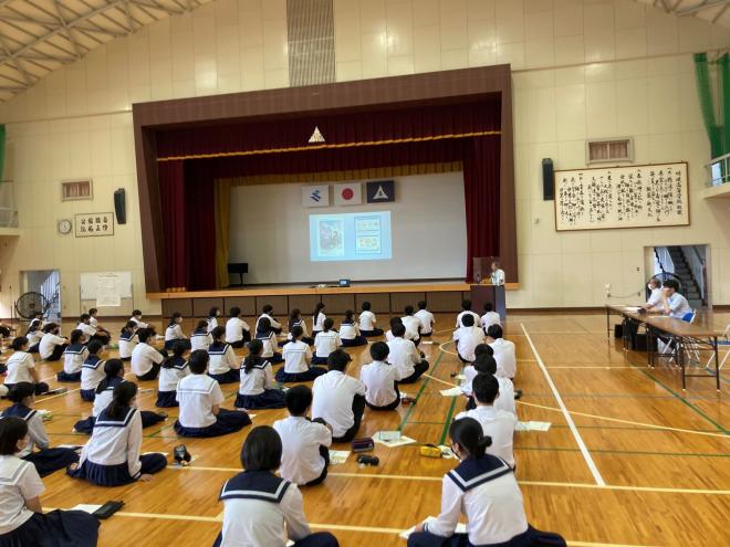 前方のスクリーンに映像が写し出され、体育館の右前方の演台で男性が話しているのを、床に体育座りをして聞いている生徒たちの後ろ姿の生徒