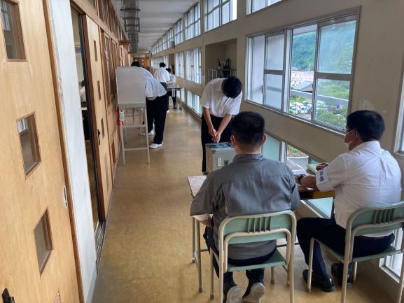 廊下に設置された投票スペースで男子生徒が投票用紙を投票箱に入れる様子を見ている先生方の写真