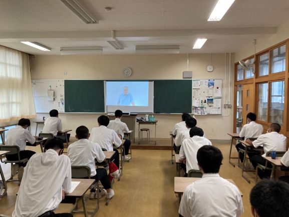 教室前方に置かれたモニターに映し出された男性の映像を見ながらリモートで話を聞いている生徒たちの写真