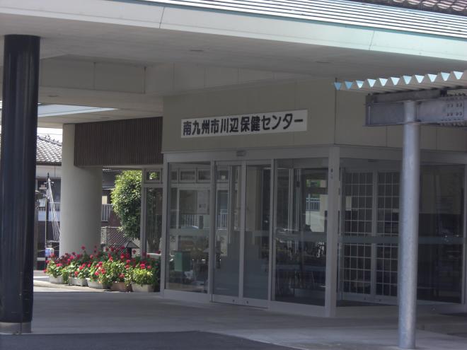 赤い花が植えらたプランタンが並べられた横にガラス張りの入口付近の南九州市川辺保健センターの写真