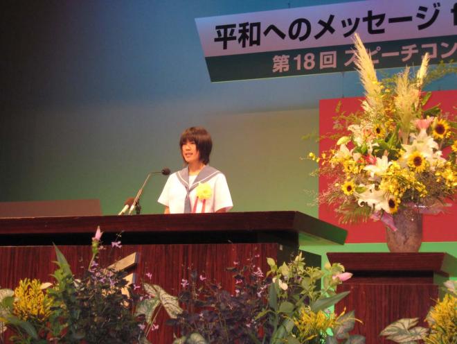 沢山の花が飾らた演壇で、セーラー服姿の中学生がスピーチをおこなっているコンテスト様子の写真