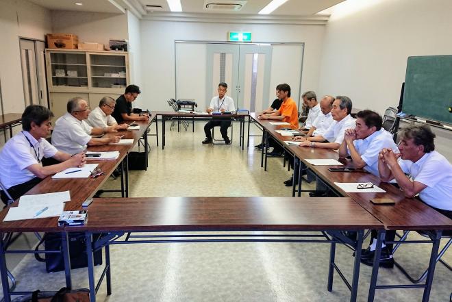右壁側に可動式の黒板が設置されている会議室内で、長机をロの字に設置した席に座っている参加者が会議を行っている様子の写真