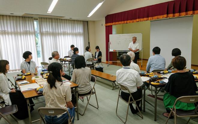 2つの長机をつけた3つのグループに分かれた席に座っている参加者が、ホワイトボードやスクリーンが設置された小さなステージ上に立っている男性の話を聞いている会議の様子の写真