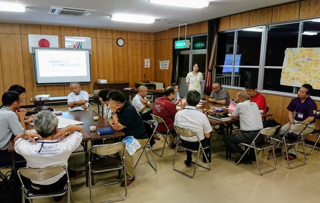 板張りで囲まれた室内で長机をつけて2つのグループに分かれて着席している参加者が、真剣にな表情で話し合いを行っている会議の様子の写真