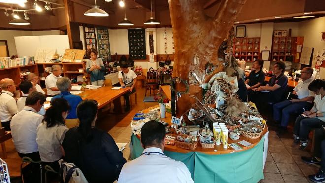中央に大きな木が立っている店内の会場で並べた椅子に座っている参加者が、左奥に立っている女性の話を聞いている会議の様子の写真