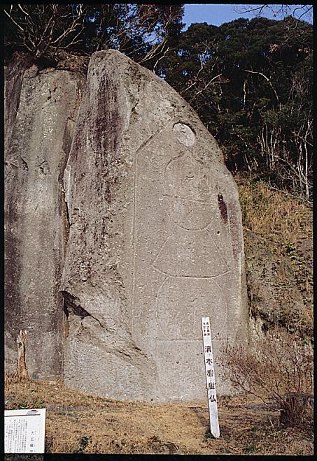 清水磨崖仏群と書かれた標柱の奥の岩壁に大きな五輪塔が刻まれている写真