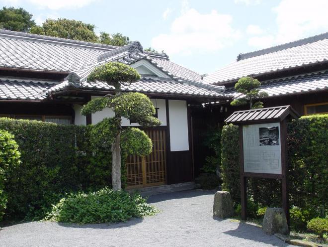 手前に案内板や生垣があり、その奥にL字形に2軒の日本家屋が並んでいる佐多直忠艇の外観写真