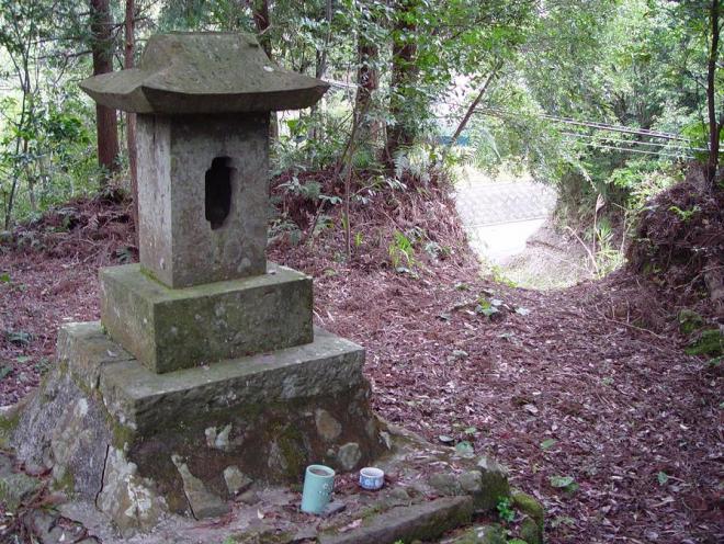 木々に囲まれた高台に、中央に楕円形の窓がある長方形の石の上に、入母屋作りの屋根を載せた祠が建っている写真