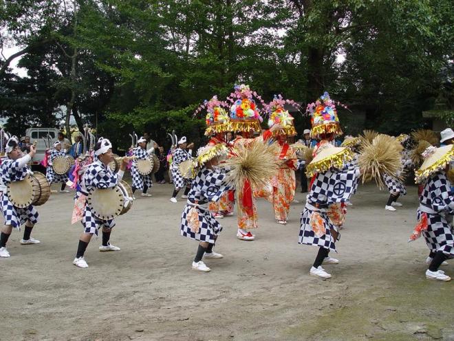 市松模様の衣装を着て刀を背負った「脇太鼓」役、藁を持った先導役の「ワラフリ」の男性たちが周囲を囲み、色鮮やかな振り袖に花がさ姿の衣装を着た「中入り」の女性たちが中央で踊っている上山田の太鼓踊りの様子の写真