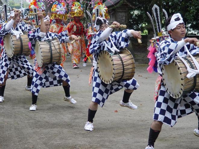 頭に鉢巻、市松模様の衣装を着て刀を背負った「脇太鼓」役の男性たちと、色鮮やかな振り袖に花がさ姿の衣装を着た「中入り」の女性たちが踊りながら境内を歩いている上山田の太鼓踊りの様子の写真