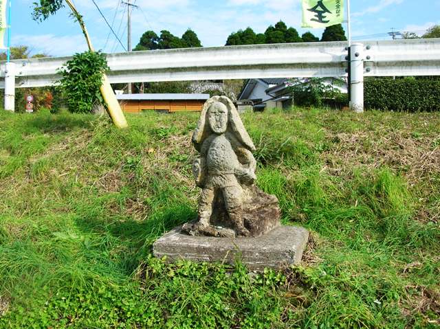 ガードレールの手前の斜面に、笠を被り,左手に鍬を持ち仁王立ちに立っている田の神様の写真