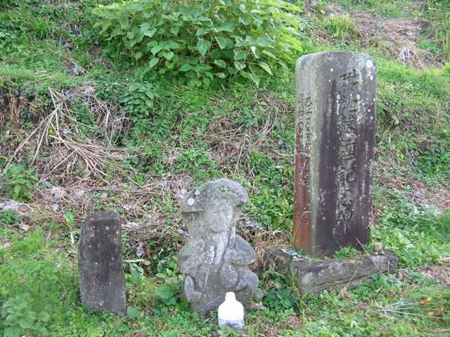 大きな記念碑と小さな石碑の間に横一列に並んで立っている、右手にオデクワ,左手にメシゲを持った田の神様の写真