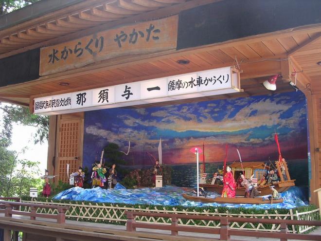水からくりやかたの木看板が設置された屋根付きの舞台上で、国選択無形民俗文化財 那須与一 薩摩の水車からくりと記載された白い横長灯籠の下、海上の船首に掲げられた扇の的を弓矢で狙っている那須与一や源頼朝達のからくり人形の写真