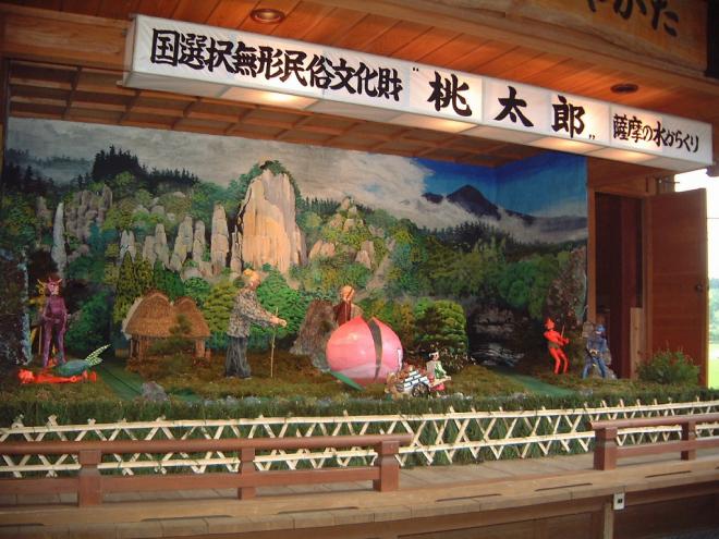 屋根付きの舞台上で、国選択無形民俗文化財 桃太郎 薩摩の水からくりと記載された白い横長の灯籠の下、舞台上で山々を背景におじいさんとおばあさん、大きな桃、桃太郎、大きな鬼などが設置されたからくり人形の写真