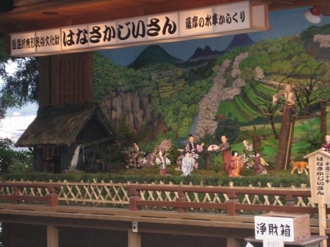屋根付きの舞台上に、国選択無形民俗文化財 はなさかじいさん 薩摩の水車からくりと記載された白い横長灯籠の下、おじいさんが枯れた木に登り花を咲かせ喜ぶ大人や子供たちのからくり人形の写真
