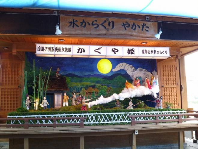 水からくりやかたの木看板が設置された屋根付きの建物内に、国選択無形民俗文化財 かぐや姫 薩摩の水からくりと記載された白い横長の灯籠の下、地上から月へと伸びる長い雲の上にかぐや姫や月からかぐや姫を迎えに来た使者を弓矢や槍で引き留めようとする人々のからくり人形の写真