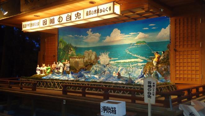 屋根付きの舞台上で、国選択無形民俗文化財 因幡の白兎 薩摩の水車からくりと記載された白い横長の灯籠の下、白いうさぎや、海の中から顔を出すたくさんのサメ、うさぎにいじわるをする大勢の神たちのからくり人形の写真