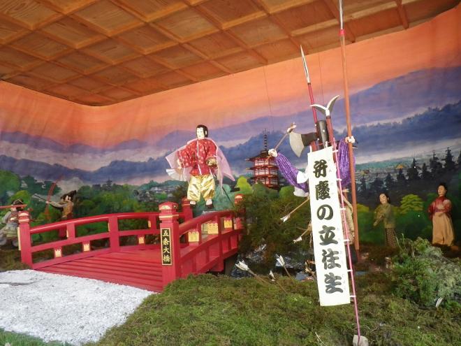 屋根付きの舞台上で、中央に設置された朱色の五条大橋をピンク色の衣を羽織った牛若丸と、手前には弁慶の立ち往生と書かれたのぼり旗と背丈以上の大刀を持った弁慶達のからくり人形の写真