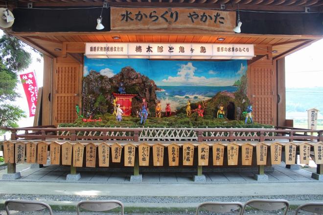 水からくりやかたの木看板が設置された屋根付きの舞台上で、国選択無形民俗文化財 桃太郎と鬼ヶ島 薩摩の水からくりと記載された白い横長の灯籠の下、刀を持った桃太郎がたくさんの赤鬼、青鬼、黄鬼と戦っている様子のからくり人形の写真
