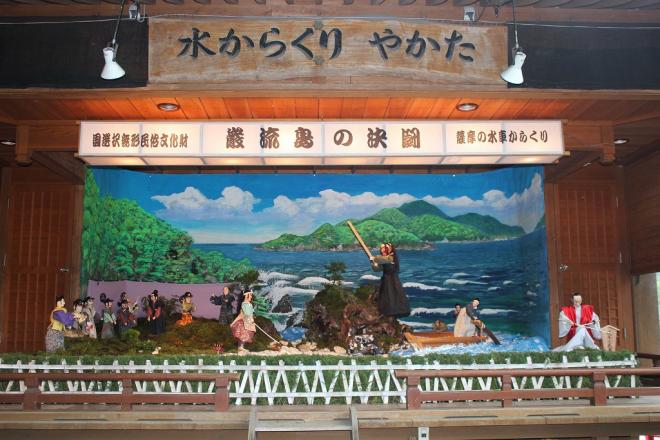 水からくりやかたの木看板が設置された屋根付きの建物内に、国選択無形民俗文化財 巌流島の決闘 薩摩の水車からくりと記載された白い横長の灯籠の下、中央には剣を持ち戦う佐々木小次郎と宮本武蔵、戦いを見守るたくさんの弟子たちのからくり人形の写真