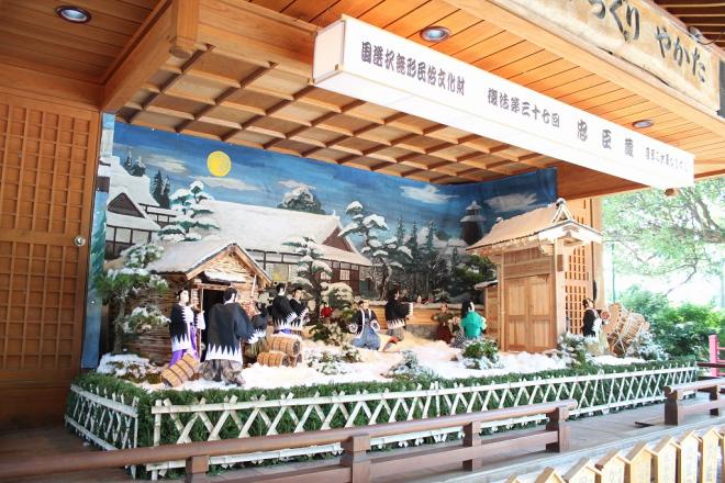 屋根付きの舞台上に、国選択無形民俗文化財 忠臣蔵 薩摩の水車からくりと記載された白い横長灯籠の下、炭小屋や立派な門が設置された吉良上野介邸で浅野内匠頭の敵を取る赤穂浪士達のからくり人形の写真
