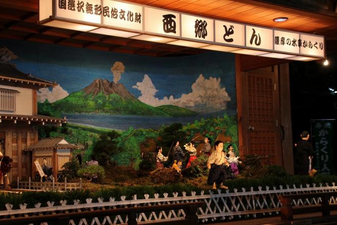 屋根付きの舞台上で、国選択無形民俗文化財 西郷どん 薩摩の水車からくりと記載された白い横長灯籠の下、刀を持った武士達が戦っている西南戦争を表したからくり人形の写真