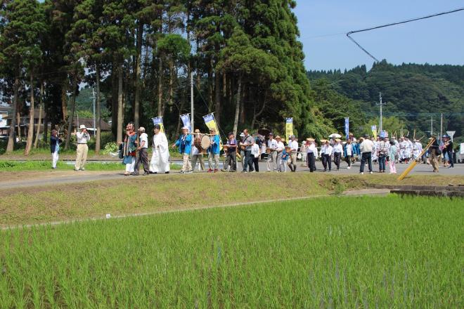 手前に水田が広がる道路にて、赤いお面の猿田彦を先頭に神職や太鼓等たくさんの参加者が列をなして御田へ移動している様子の写真