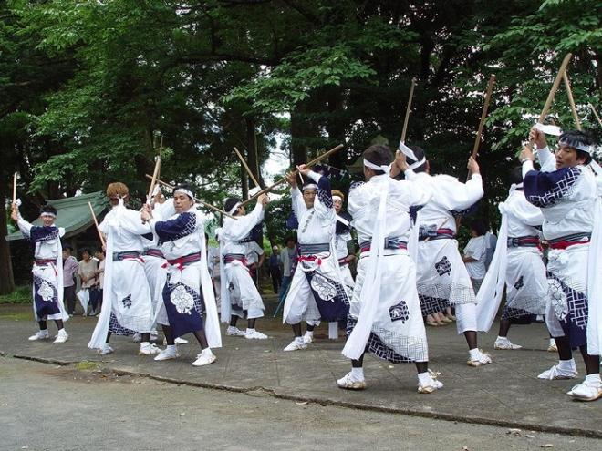 飯倉神社境内の木々が立ち並ぶ参道にて、揃いの白い衣装に身を包んだ白いハチマキ姿の男性陣が3列縦隊で六尺棒と三尺棒を使い舞を披露している松崎棒踊りの写真