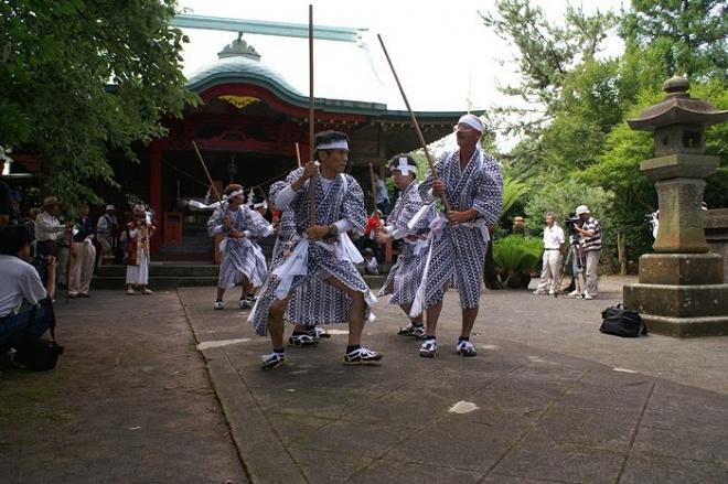 朱色の飯倉神社を背景に、両側に見学客が見守る中、神社中央の参道を白いハチマキ姿で揃いの衣装姿の踊り手が2列に並び六尺棒を持ち舞を披露する宮小路棒踊りの写真