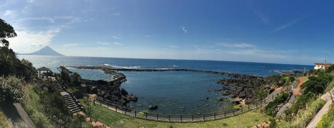 海の向こうに開聞岳を眺める番所鼻自然公園にて、環状型の岩礁を緩やかな地平線と共に高台から撮影されたパノラマ写真
