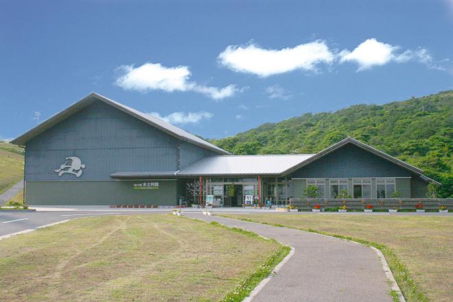 白い雲が浮かぶ青空の下、後方には小高い山があり、広々とした場所にあるグレーの外壁をした三角屋根の畑の郷水土利館の外観写真