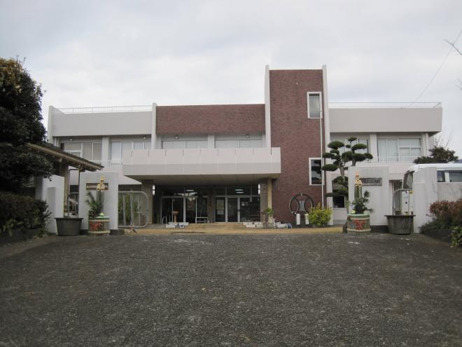 白色と一部茶色の外壁をした2階建ての建物で白い門の両側に門松が置かれている郡地区公民館の外観写真