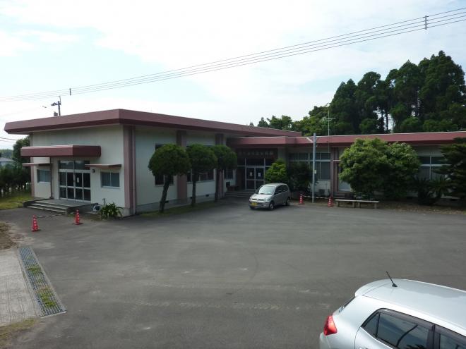 コンクリートで舗装された敷地奥に建つ、白壁で柱や屋根が薄ピンク色のL字型平屋建ての松ヶ浦地区公民館の建物外観の写真