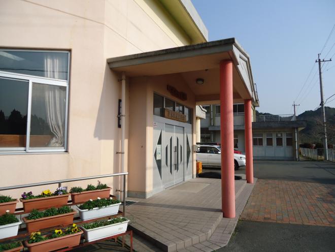 ベージュ色の外壁で2本の赤い円柱の柱で支えた庇がある清水地区公民館の入り口を、花が植えられたプランターが並んでいる左側から撮影した写真