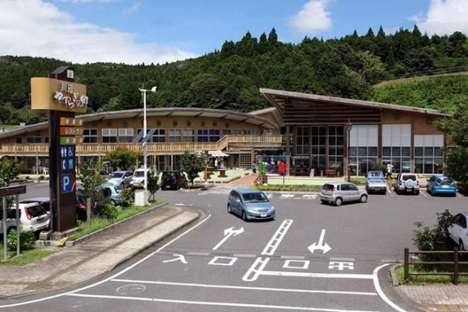 道路沿いに「川辺やすらぎの郷」と書かれた看板があり、広々とした駐車場と丸みを帯びた屋根の建物と軒が三角に突き出した形の屋根をした建物が隣接している川辺やすらぎの郷の外観写真