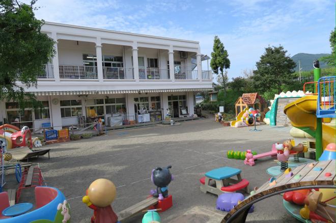 木々に囲まれた広い園庭に沢山の遊具が設置されている、白壁で2階建ての南九州勝縁こども園の写真