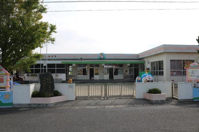 園庭の左側に大きな木や遊具があり、白壁で横長のちどり保育園の建物外観を門の外側から撮影した写真