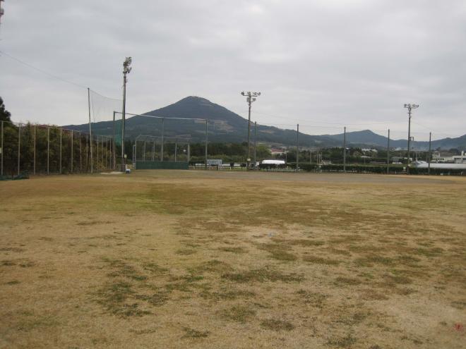 背景に大きな山がそびえ、木立やフェンスで囲まれ、夜間照明が設置されている広々とした南九州市頴娃グリーン広場(ソフト)の写真