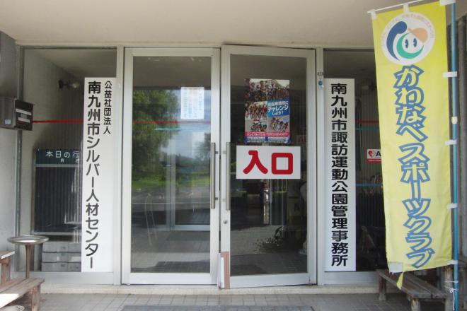 両開きの玄関で、入口と書かれたガラス窓に、南九州市諏訪運動公園管理事務所、公益社団法人南九州市シルバー人材センターの看板、かわなべスポーツクラブののぼり旗が設置された玄関前の写真