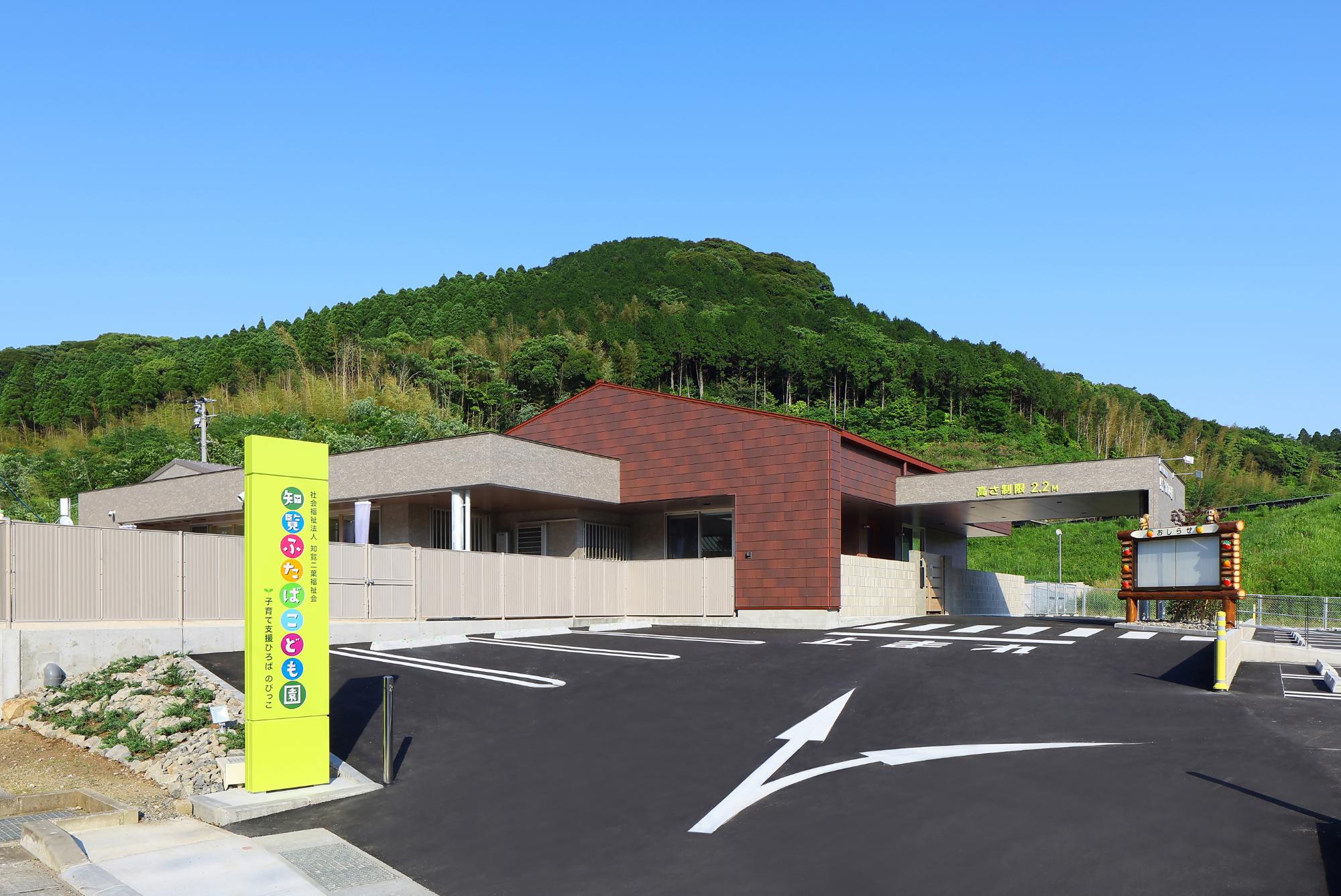 広い園庭の砂場で園児たちが遊んでおり、赤色の屋根で白壁の1階建てと2階建ての建物が併設されている大心寺二葉保育園の写真