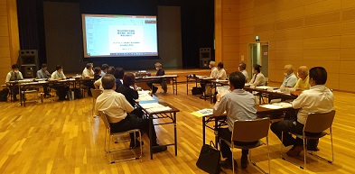 広いホールに長机をいくつか並べて計20名程が座り、ステージ上に設置されたスクリーンに映る資料を見ている第2回検討委員会の様子の写真