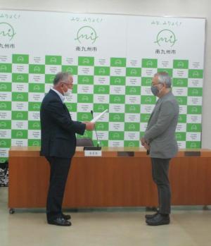会議室内のバックパネルの前で、市長が向かい合って立つ男性に手に持つ詰問書の内容を読み上げている様子の写真
