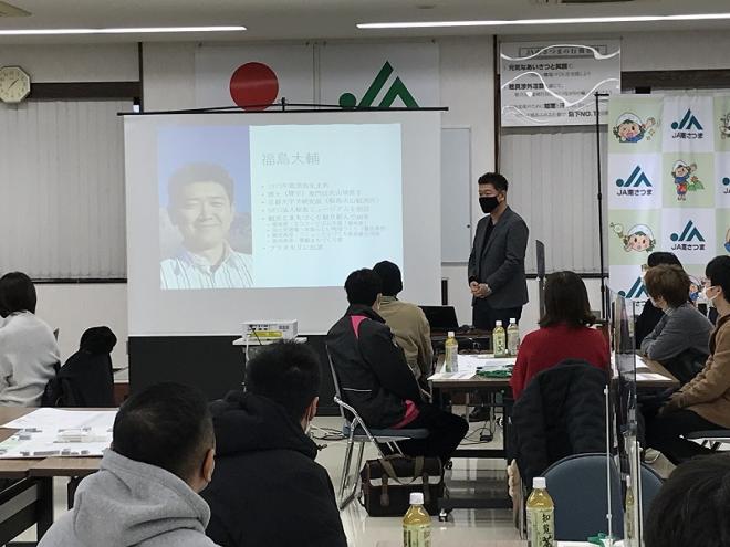 スクリーンに福島大輔さんの紹介が映し出され、スクリーンの横で男性が話をするのを座って聞いている参加者の写真