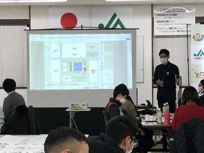 スクリーンに資料が映し出され、スクリーンの横で男性が話をするのを座って聞いている参加者の写真