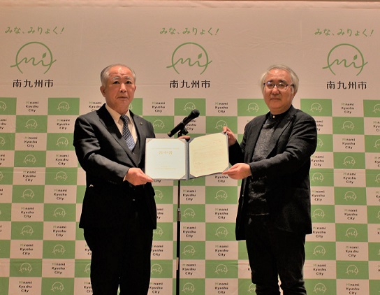 南九州市のバックパネルを背景に鰺坂委員長と塗木市長が見開きにした答申書を2人で持っている記念写真