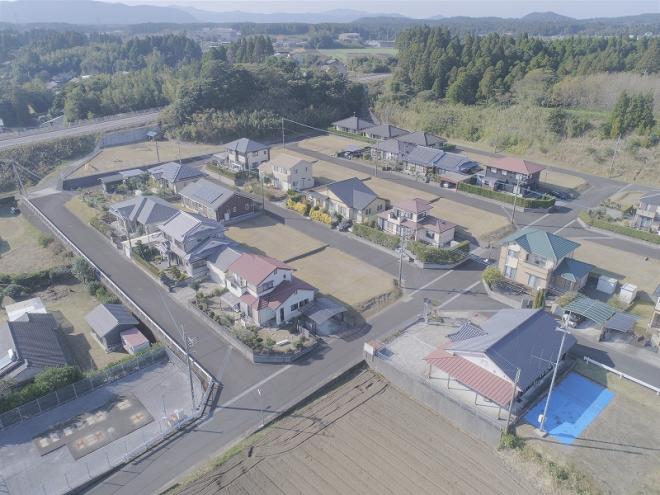 奥に山脈が広がり、赤・緑・グレーの屋根の家が建ち並んでいる団地を上空から撮影した写真