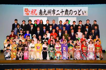 「祝南九州市二十歳のつどい」と書かれた横断幕が掲げられた舞台の上に、色とりどりの振袖を着た女性と、袴やスーツを着た男性の新成人が5列に並んで写した記念写真