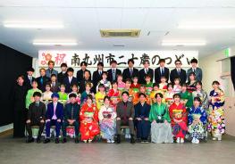 「祝南九州市二十歳のつどい」と書かれた横断幕が掲げられ会場の壁の前で、色とりどりの振袖を着た女性と、袴やスーツを着た男性の新成人が4列に並んで写した記念写真